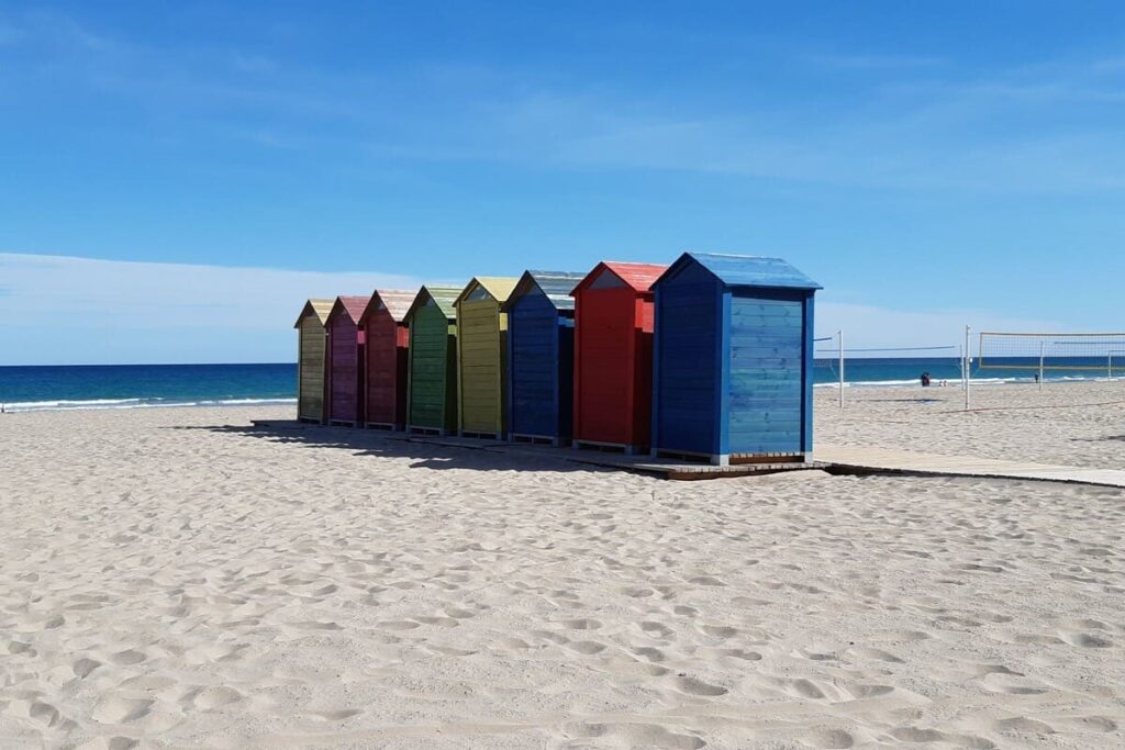 Wooden beach huts in vibrant colors next to volleyball courts on San Juan Beach, Alicante, under bright winter sun and blue sky