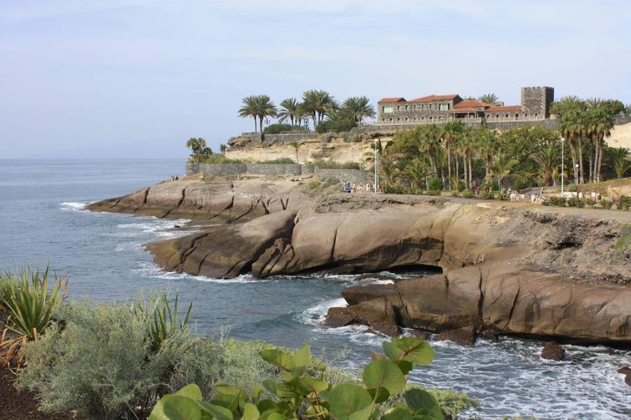Oceanfront view of Tenerife with rocky cliffs, palm trees and a historic-looking stone building